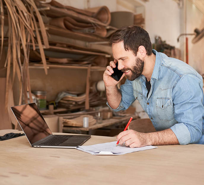 Entrepreneur dans son atelier d'artisanat avec ordinateur et smartphone qui gère son activité avec le sourire.
